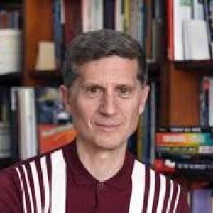 Headshot of man in front of full bookshelf wearing deep red shirt with vertical stripes