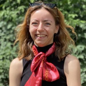 Image of smiling younger woman standing outside in front of tree wearing sleevelss black top with red scarf