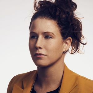 headshot of a serious woman with brown hair up in a bun wearing a black grapchic tee and deep yellow blazer