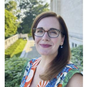 Image of smiling woman with long auburn hair and glasses standing outside wearing a floral top