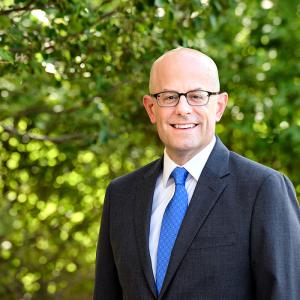 Image of man in suit with glasses standing outside in front of trees