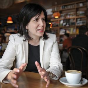 Color image of women sitting at a table with a coffee cup in front of her.  She has shoulder length brown hair, is wearing a white blazer, and appears to be in conversation.