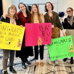 Group shot of Professor Gertsman surrounded by 4 female students with colored signs