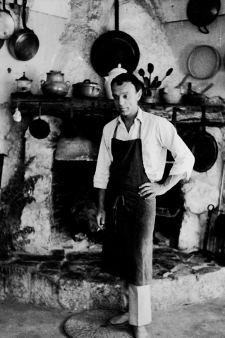 Black and white photo of white man in his thirties wearing an apron and standing in front of a kitchen fireplace with lost of pots hanging