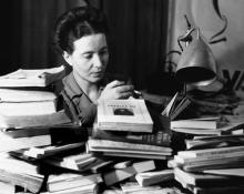 Black and white photo of Simone de Beauvoir at a desk surrounded by books