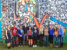 Color photo of group of diverse college students in front of a colorful outdoor mural