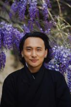 Head shot of Bhutanese man wearing black robe and blue earrings standing outside in front of flowering purple trees
