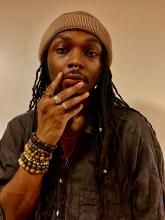 color image of African American man with long hair, wearing a beige knit cap, with his hand on his face.  He has on several beaded bracelets and has a septum ring.