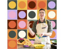 White man with dark hair and glasses with various kitchen tools wearing an apron in front of a colorful background of various circles