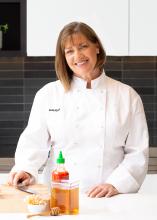 Smiling white woman with shoulder length brown hair wearing a chef's coat standing at a table