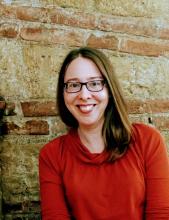 Image of smiling white woman with long brown hair, glasses, and a red sweater standing outside in front of a brick wall