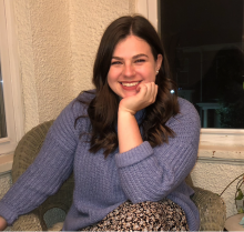 Smiling woman with long brown hair sitting in a chair resting her hand on her chin,