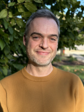 Headshot of white man wearing a dark yellow shirt standing outside in front of a tree