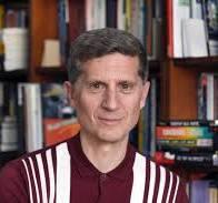 Headshot of man in front of full bookshelf wearing deep red shirt with vertical stripes