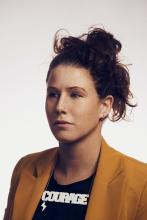 headshot of a serious woman with brown hair up in a bun wearing a black grapchic tee and deep yellow blazer