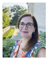 Image of smiling woman with long auburn hair and glasses standing outside wearing a floral top