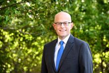 Image of man in suit with glasses standing outside in front of trees