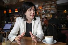 Color image of women sitting at a table with a coffee cup in front of her.  She has shoulder length brown hair, is wearing a white blazer, and appears to be in conversation.