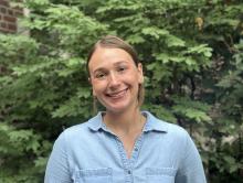 Image of smiling woman with hair pulled back wearing a blue shirt and standing outside in front of leafy trees