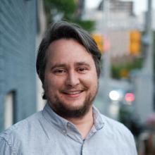 Head shot of smiling man in front of buildings with facial hair and a blue shirt