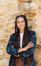 Color photo of smiling woman with long braids and arms crossed standing in front of a stone wall