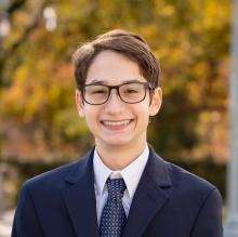 head shot of smiling younger man outside in front of autumn trees wearing a suit and tie and glasses