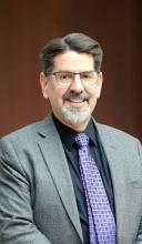 Image of smiling man with glasses and facial hair wearing a grey suit and purplish tie