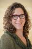 Head shot of smiling woman with shoulder length brown curly hair and glasses wearing a green jacket