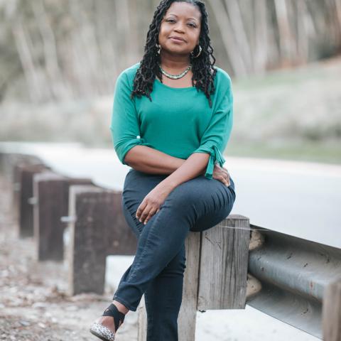 Image of African American woman with long braided hair wearing a green sweater and sitting on a guardrail