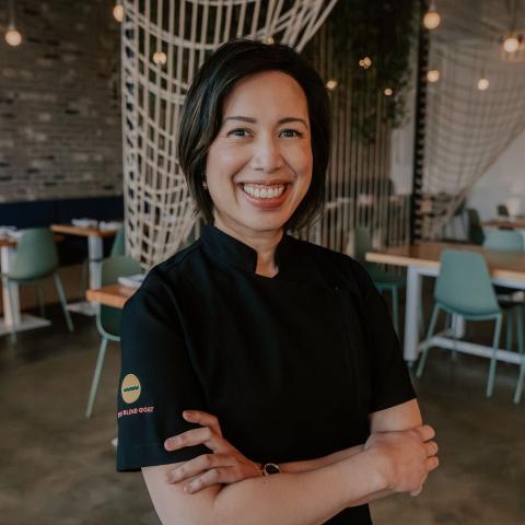 Image if smiling asian woman inside of a restaurant with arms crossed wearing black chef's jacket