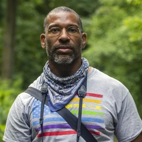 Image of African American Man outside in front of trees wearing a tee-shirt and bandana