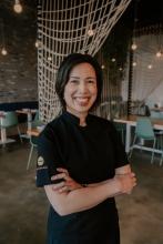 Image if smiling asian woman inside of a restaurant with arms crossed wearing black chef's jacket