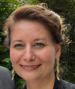 A brunette woman with long earrings smiling against a backdrop of foliage