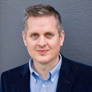 Middle-aged man with blue button up and black jacket in front of a gray background.
