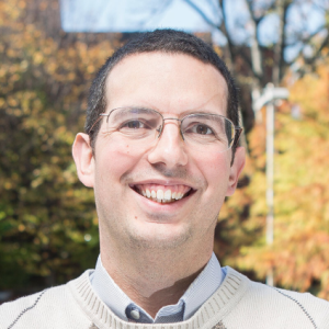 A smiling man with black hair and glasses against a background of fall trees