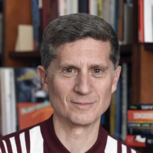 A middle-aged man in a maroon shirt sitting in front of a bookshelf