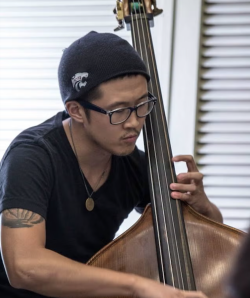 A man in a black T-shirt, glasses, and a hat plays the upright bass against a white background