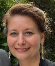 A brunette woman with long earrings smiling against a backdrop of foliage