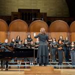 Group of female singers onstage in black, with a piano on the left and conductor in front of the group.