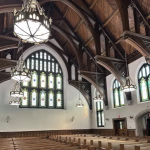 The arched ceiling of a chapel with hanging lamps in the foreground and a stained-glass window in the background 