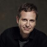 A man with sandy brown hair, a black dress shirt, and a tie looking directly at the camera against a grey background