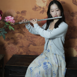 Erin Huang, young female student with dark hair wearing dress with blue flowers, playing flute.