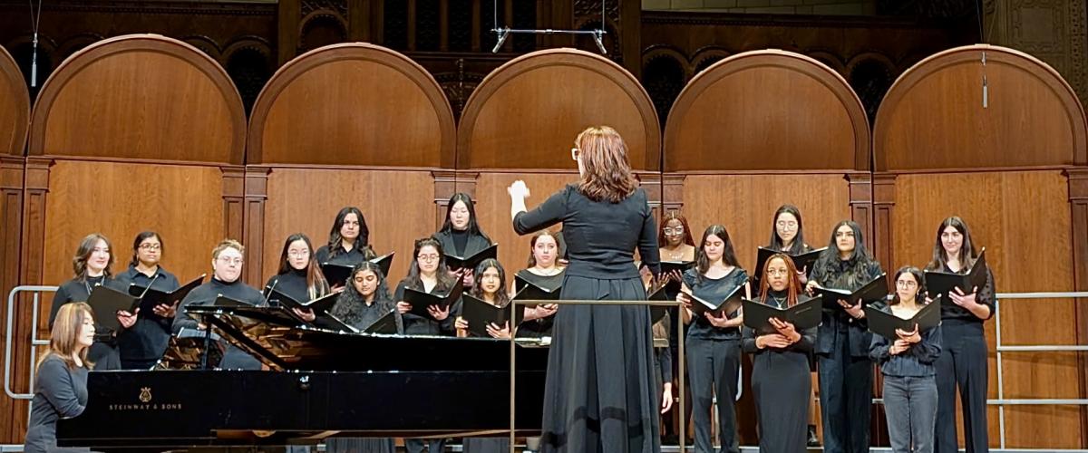 Group of female singers onstage in black, with a piano on the left and conductor in front of the group.