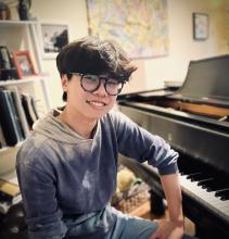 Austin Lu, youn male student with dark hair wearing gray shirt, sitting on piano bench facing camera