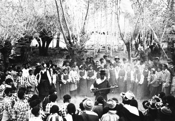 Black & white photo of a group of children gathering around a musician in robes in a grove of trees