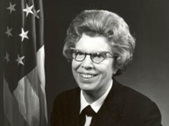Alene Duerk smiles at camera while wearing glasses and a military uniform next to an American flag
