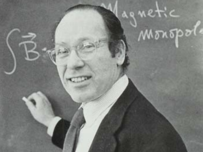 Stefan Machlup writes at a blackboard while wearing formal attire and smiling at the camera