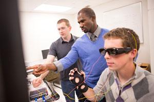 Two professors observe a student wearing goggles and a glove hooked up to a machine