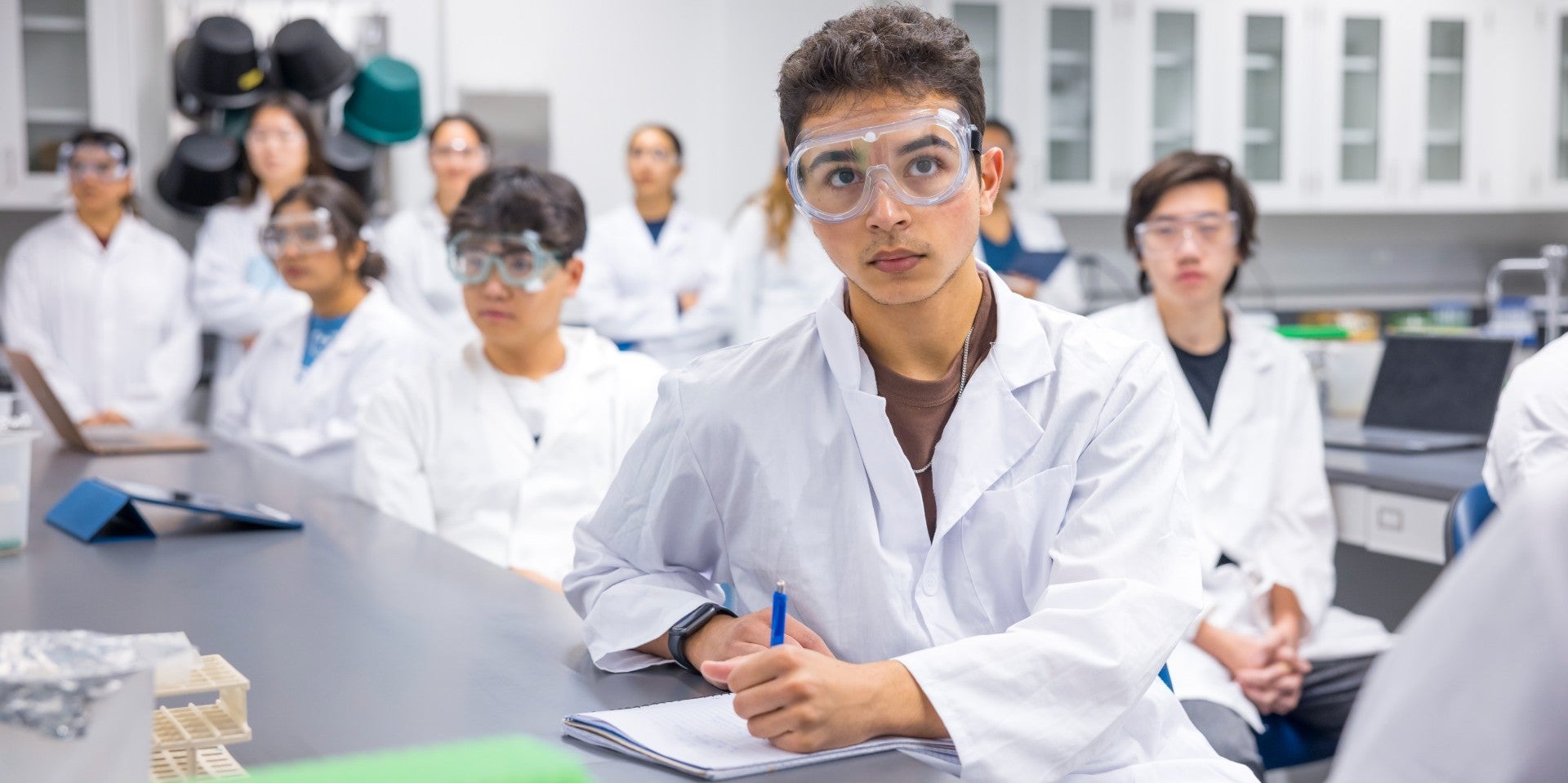 Students in a labratory setting wear PPE while taking notes.