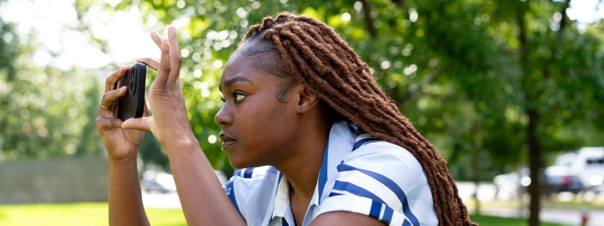 A student focuses intently on her phone screen while taking a picture outdoors.